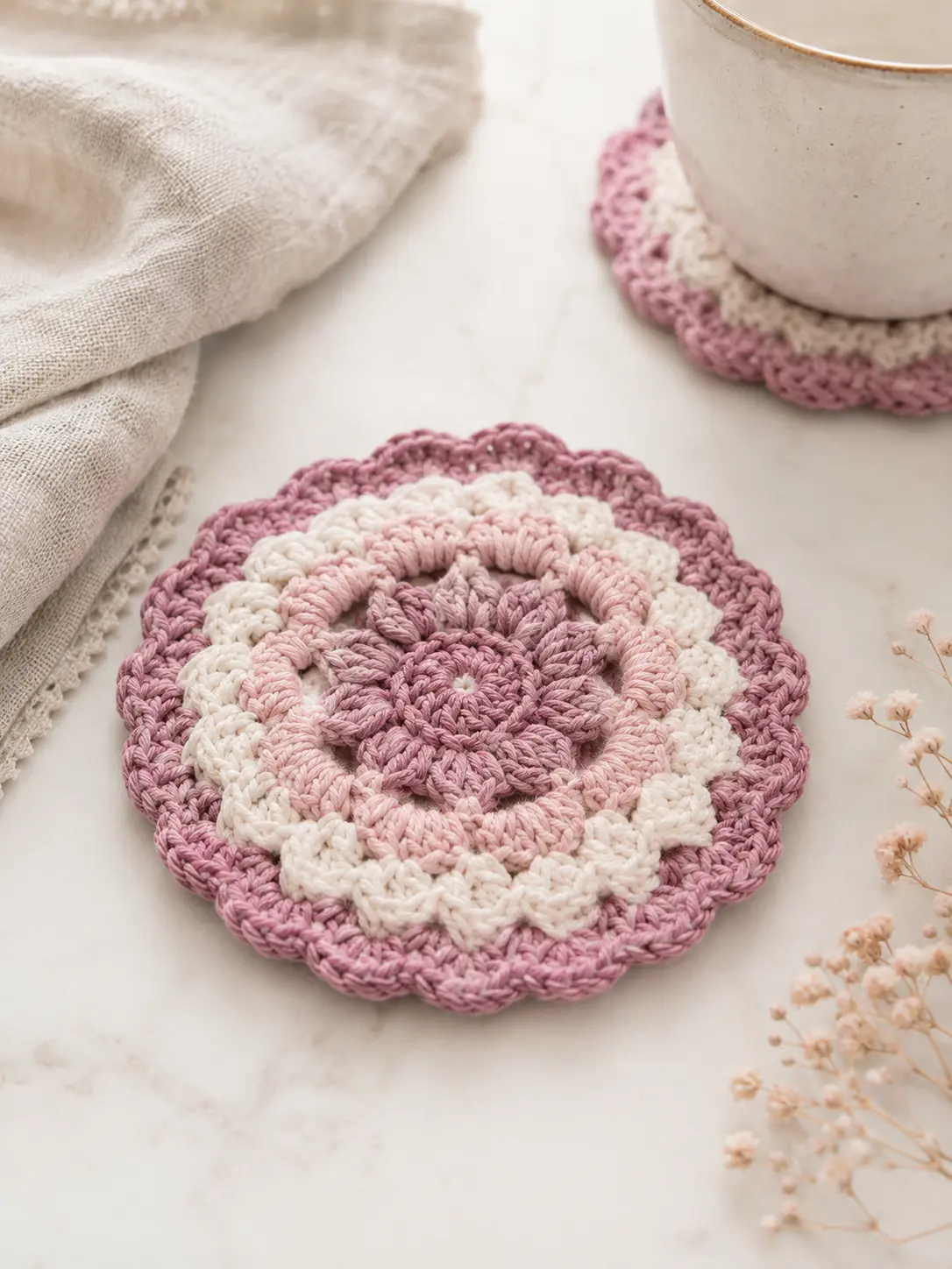 Posavasos redondo de crochet en tonos malva, rosa suave y crema sobre una mesa clara, con una taza al fondo y detalles delicados alrededor.